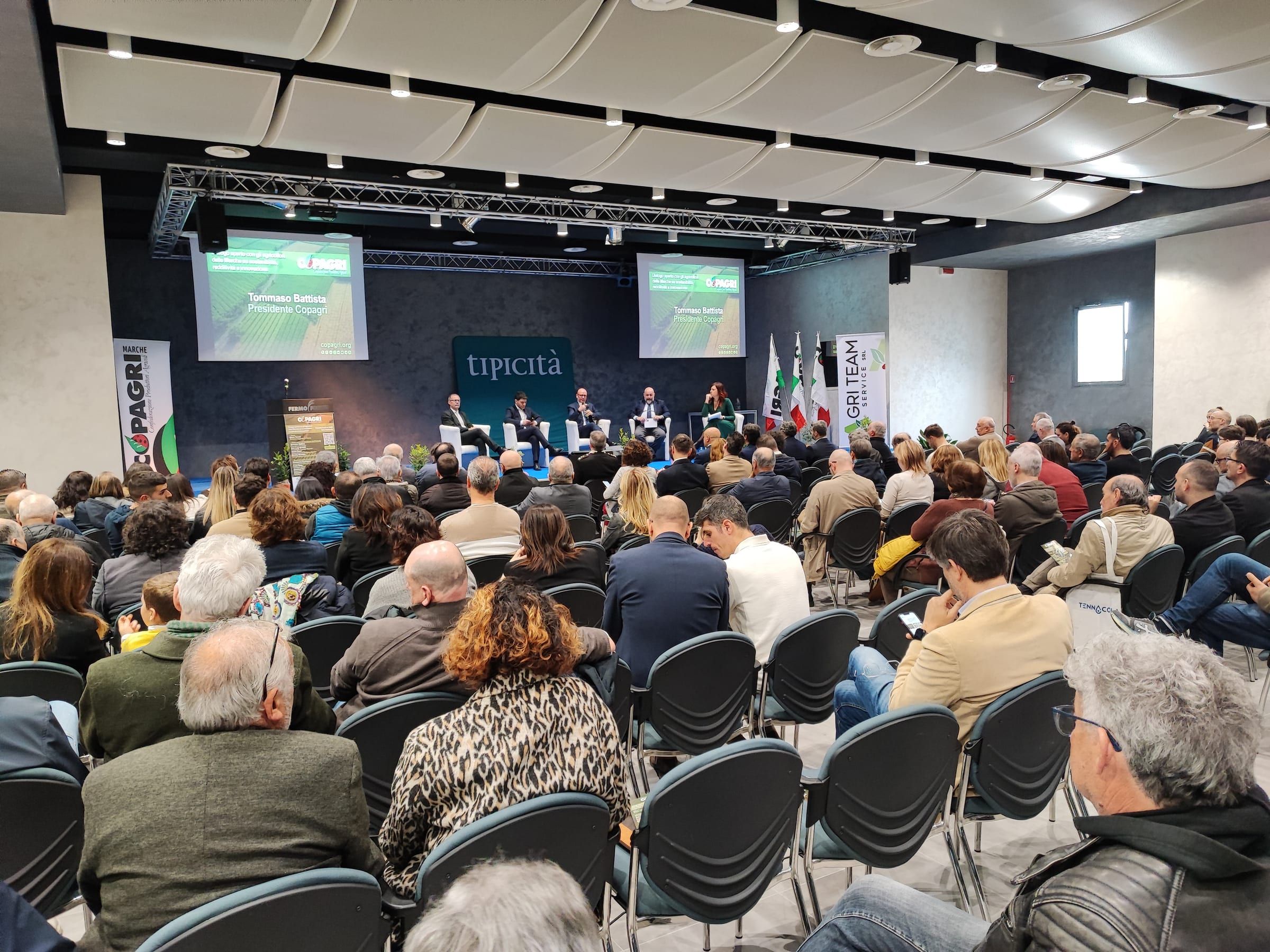 Una sala convegni piena, ripresa dal fondo: file di persone sedute davanti al palco e allo schermo con il marchio Tipicità.