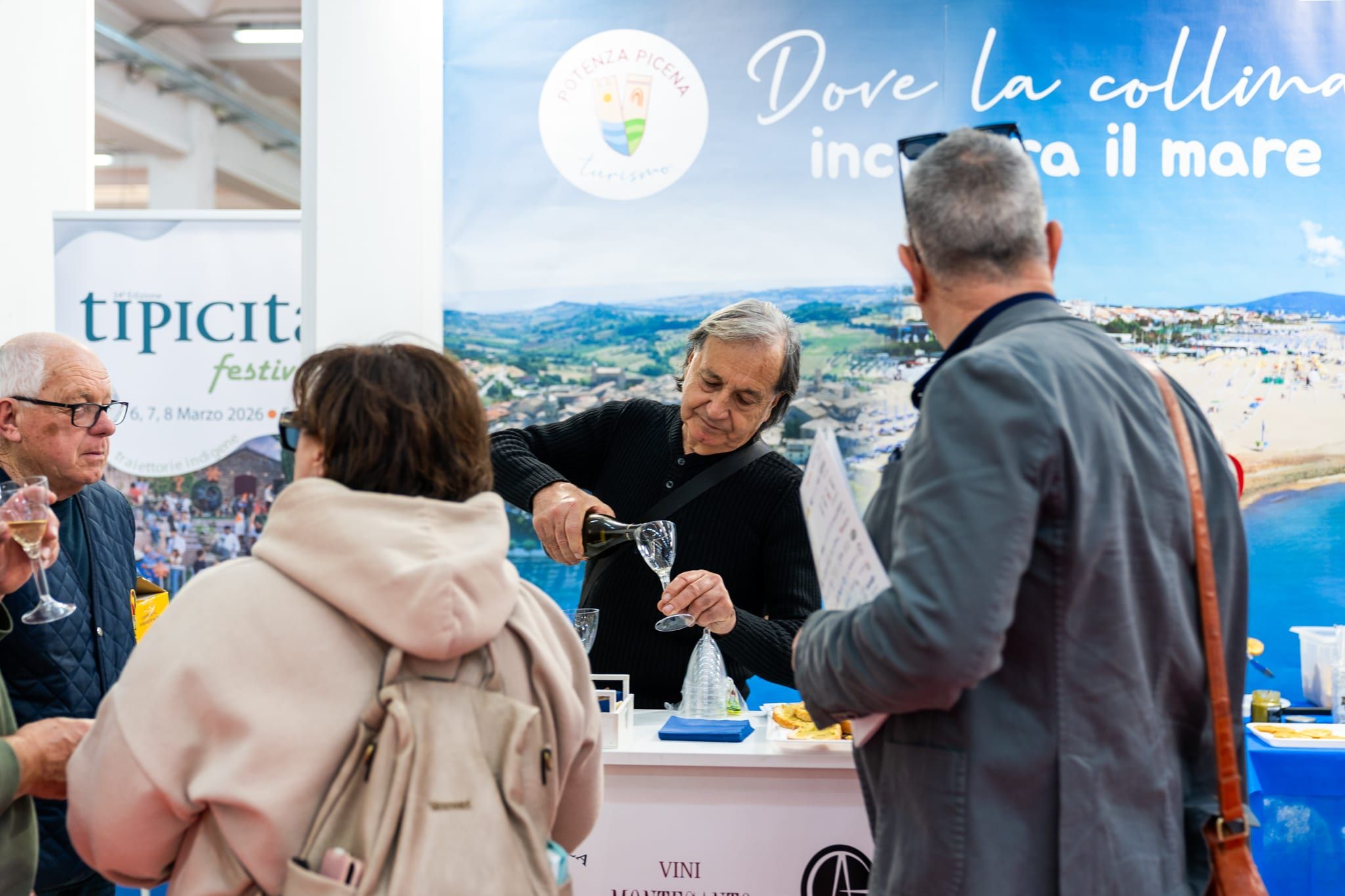 A uno stand di territorio, sotto una grande fotografia di colline che scendono sul mare, un produttore versa il vino a due visitatori fermi a parlare con lui.