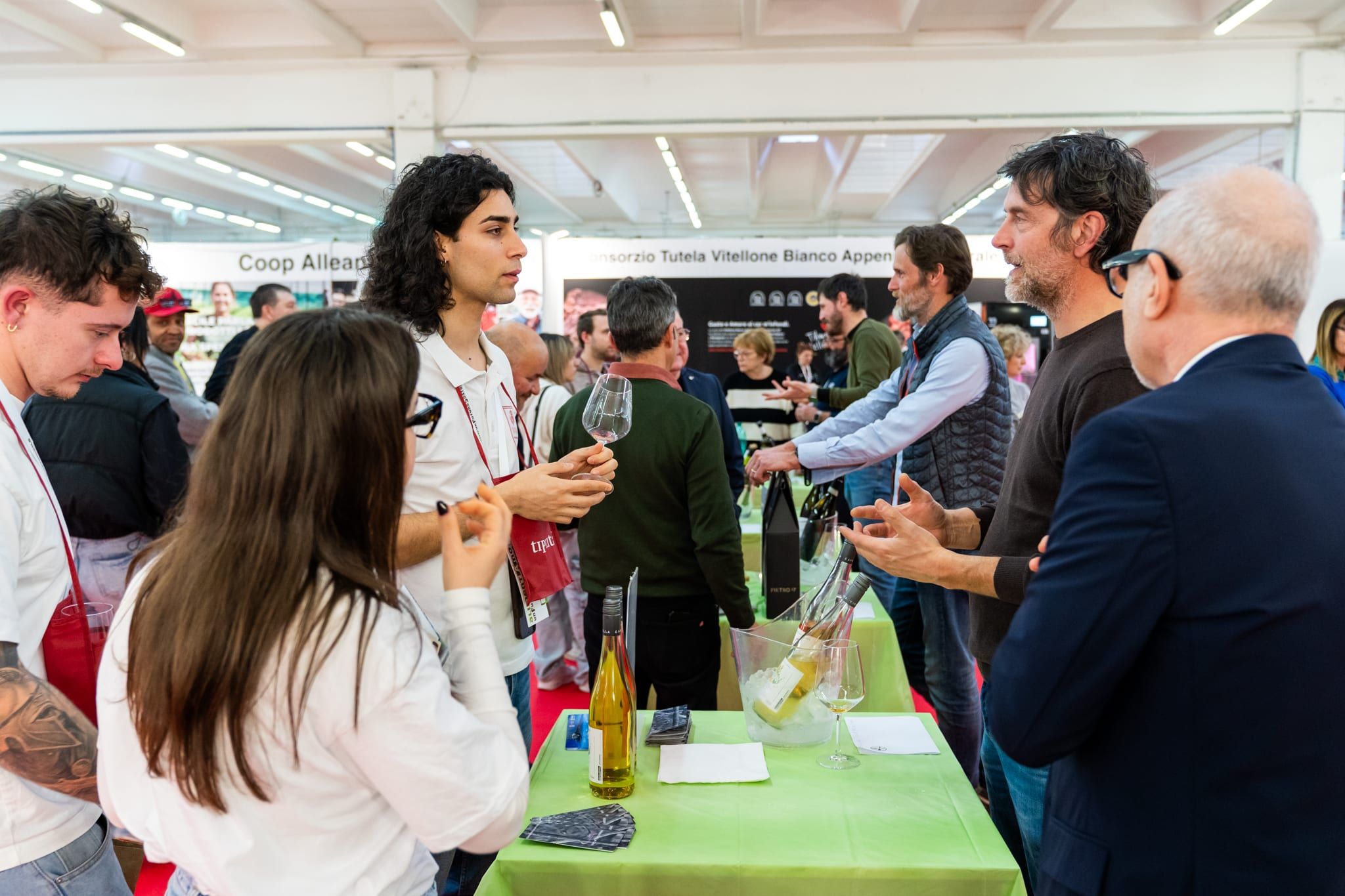 Capannelli di persone in piedi fra due stand, calici in mano, che parlano davanti ai tavoli verdi della degustazione.