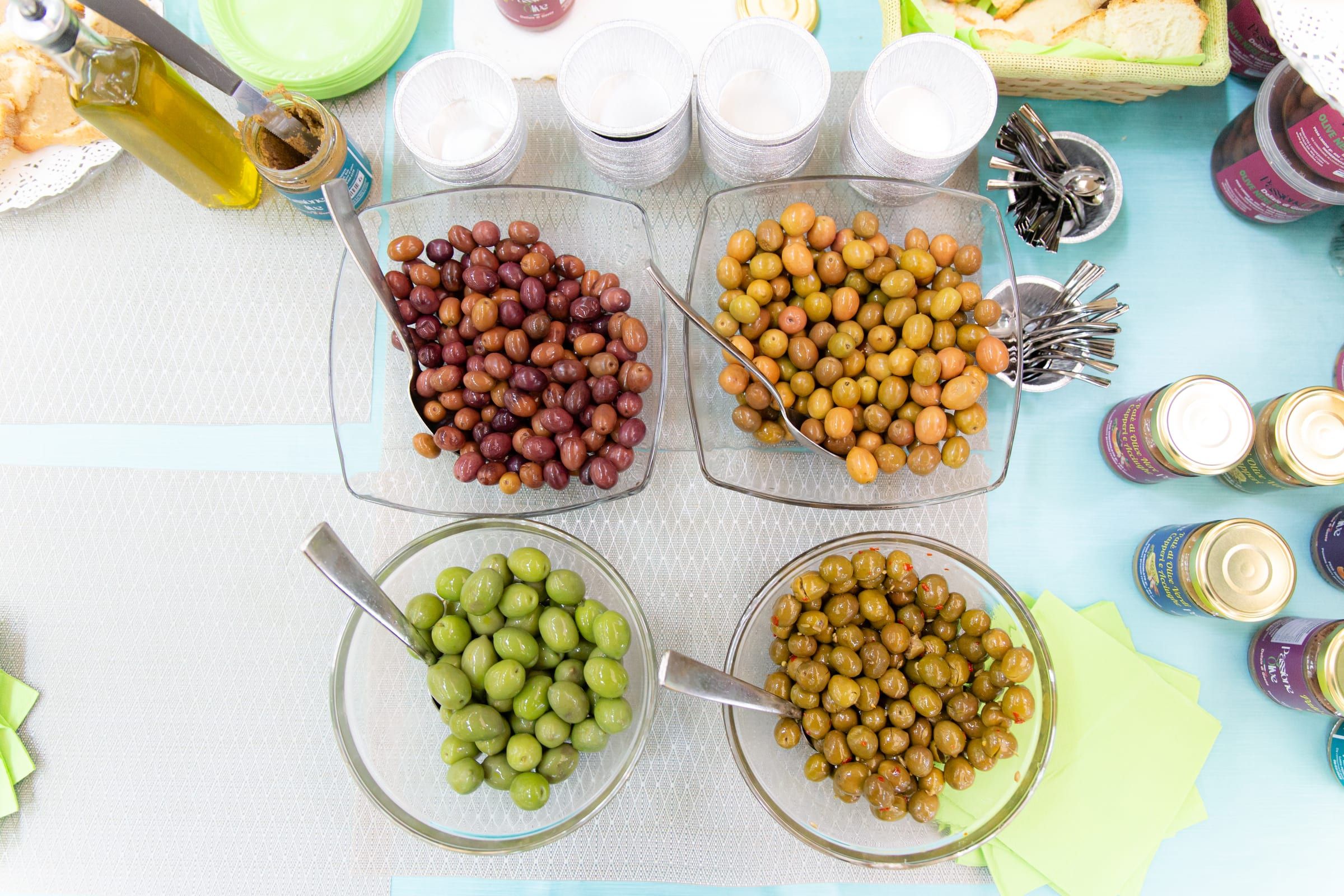 Quattro ciotole di vetro viste dall'alto, con olive verdi, dorate, rosse e scure, i cucchiai appoggiati dentro e una bottiglia d'olio di fianco.